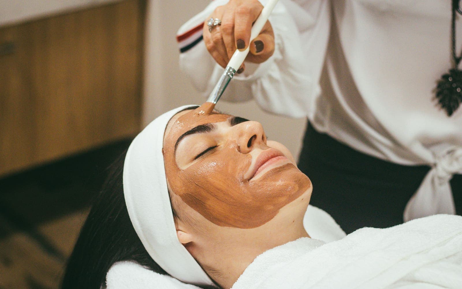 A therapist brushing a clay treatment mask onto a client's face during a professional facial