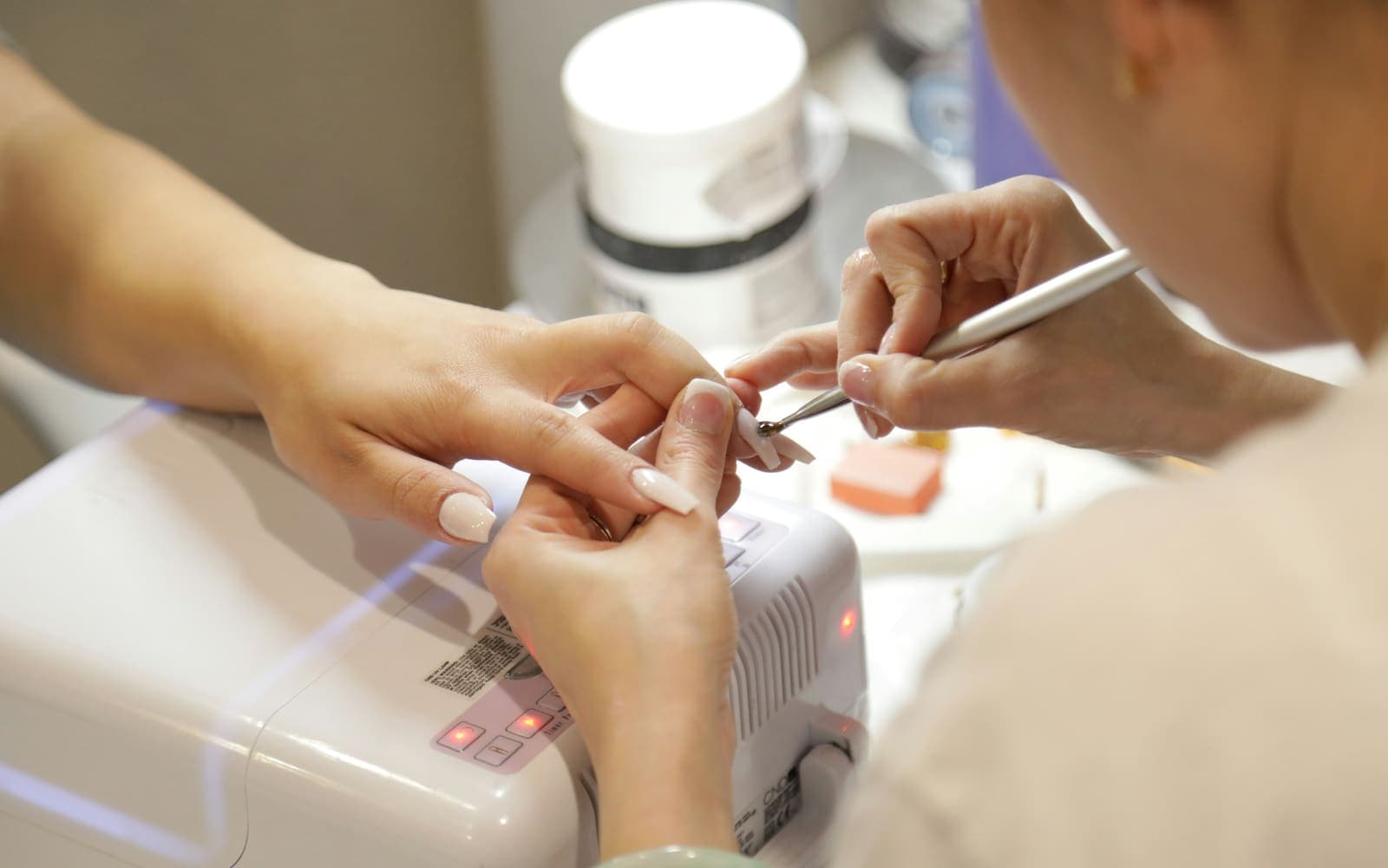 A nail technician shaping a client's nail at the desk beside a curing lamp