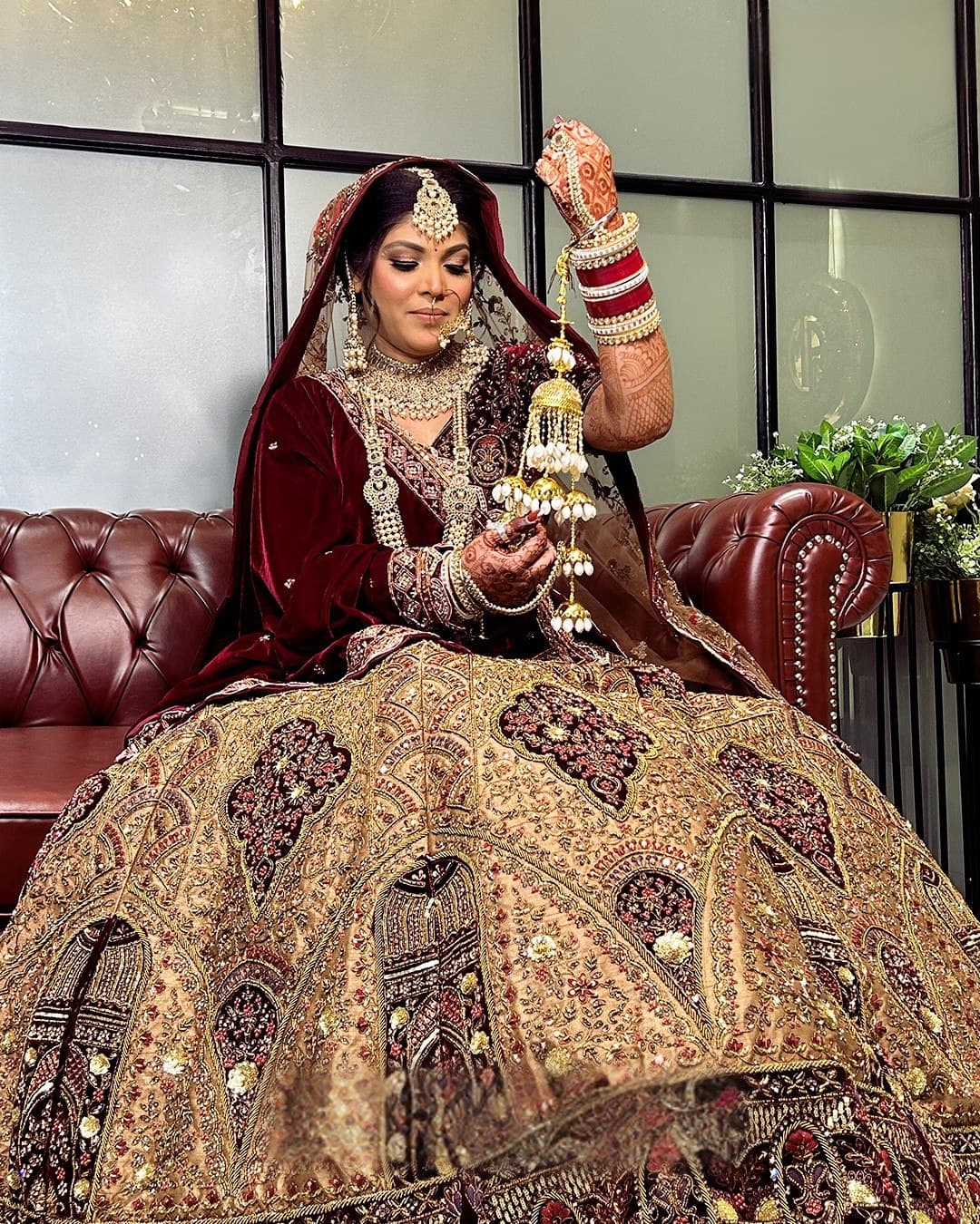 A bride in a maroon and gold lehenga seated with her kaleere raised