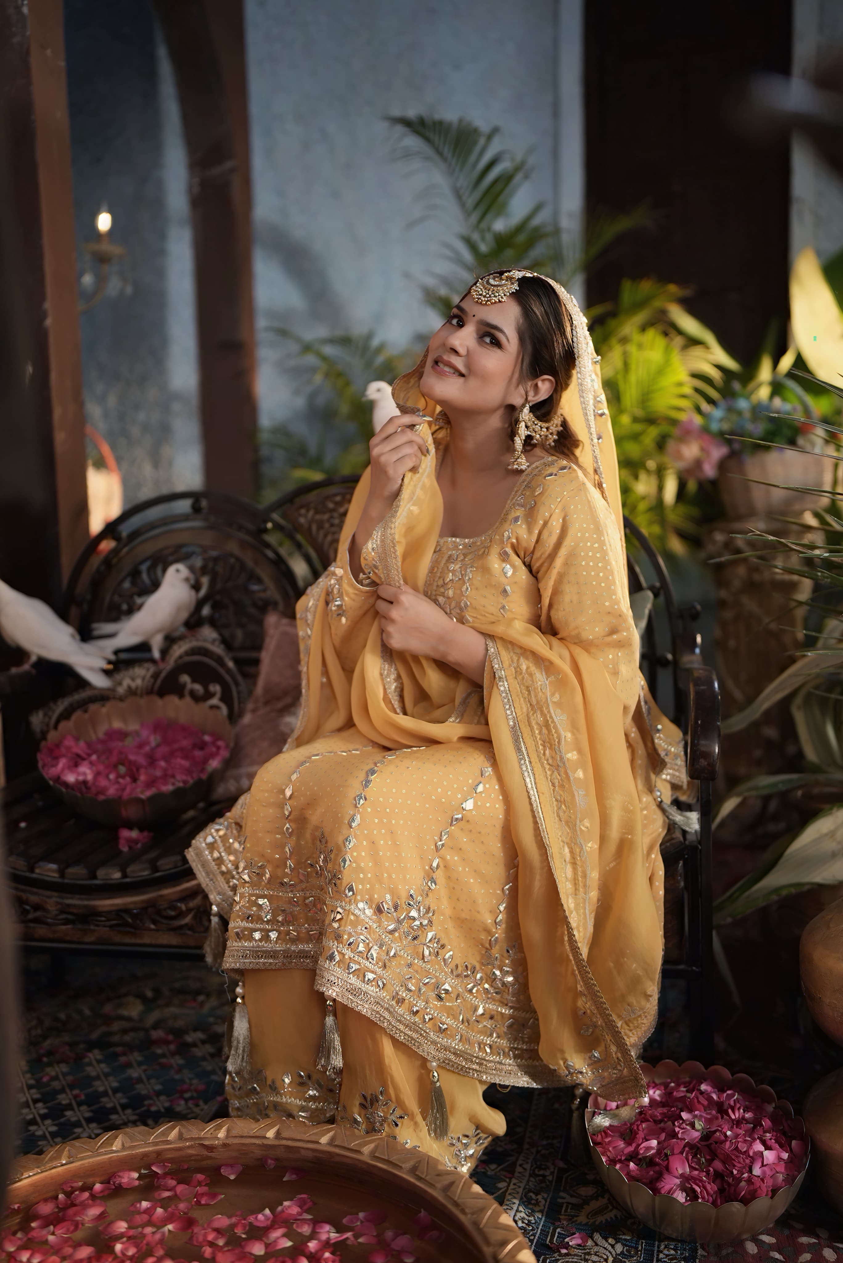 A celebrity client styled by Anthéa Makeover in a gold haldi look, seated with doves and rose petals
