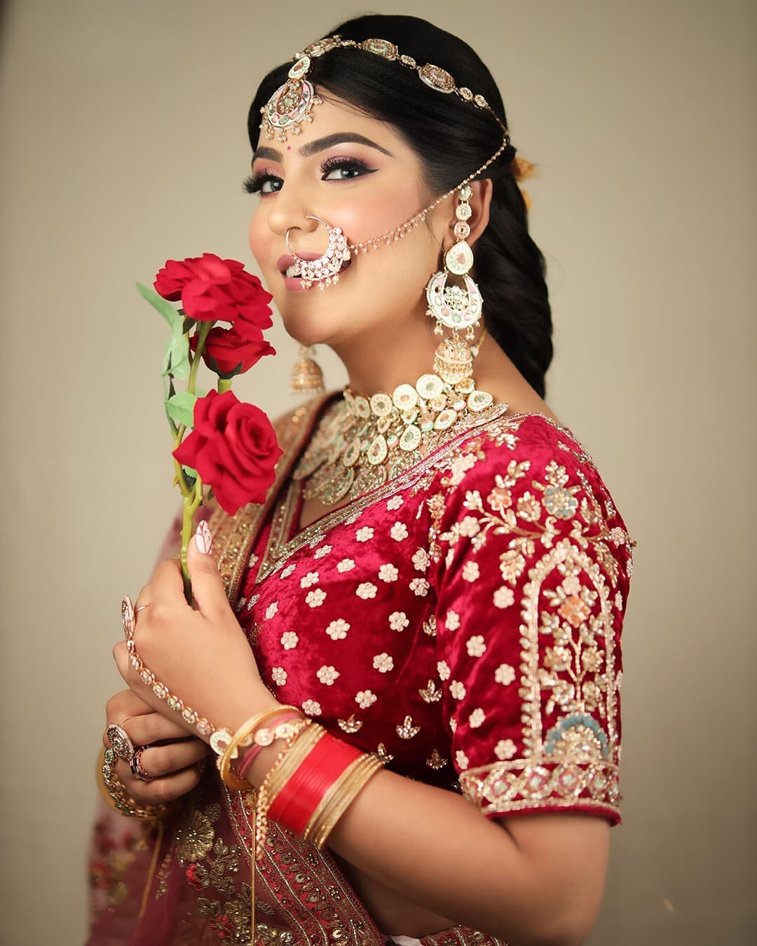 A bride in a red velvet blouse holding red roses, in a matha patti and nath